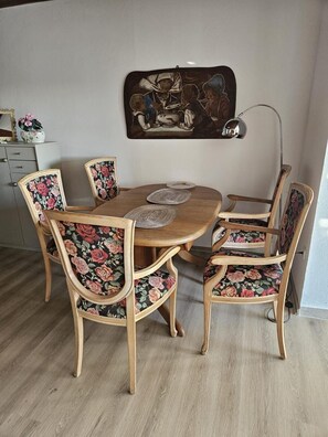 Uma área de refeições com mesa de madeira e cadeiras estofadas com padrão floral, um objeto decorativo na parede acima e uma luminária à direita.