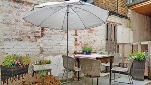 Eine Terrasse mit Korbstühlen, einem Tisch, einem weißen Sonnenschirm und Kübelpflanzen.