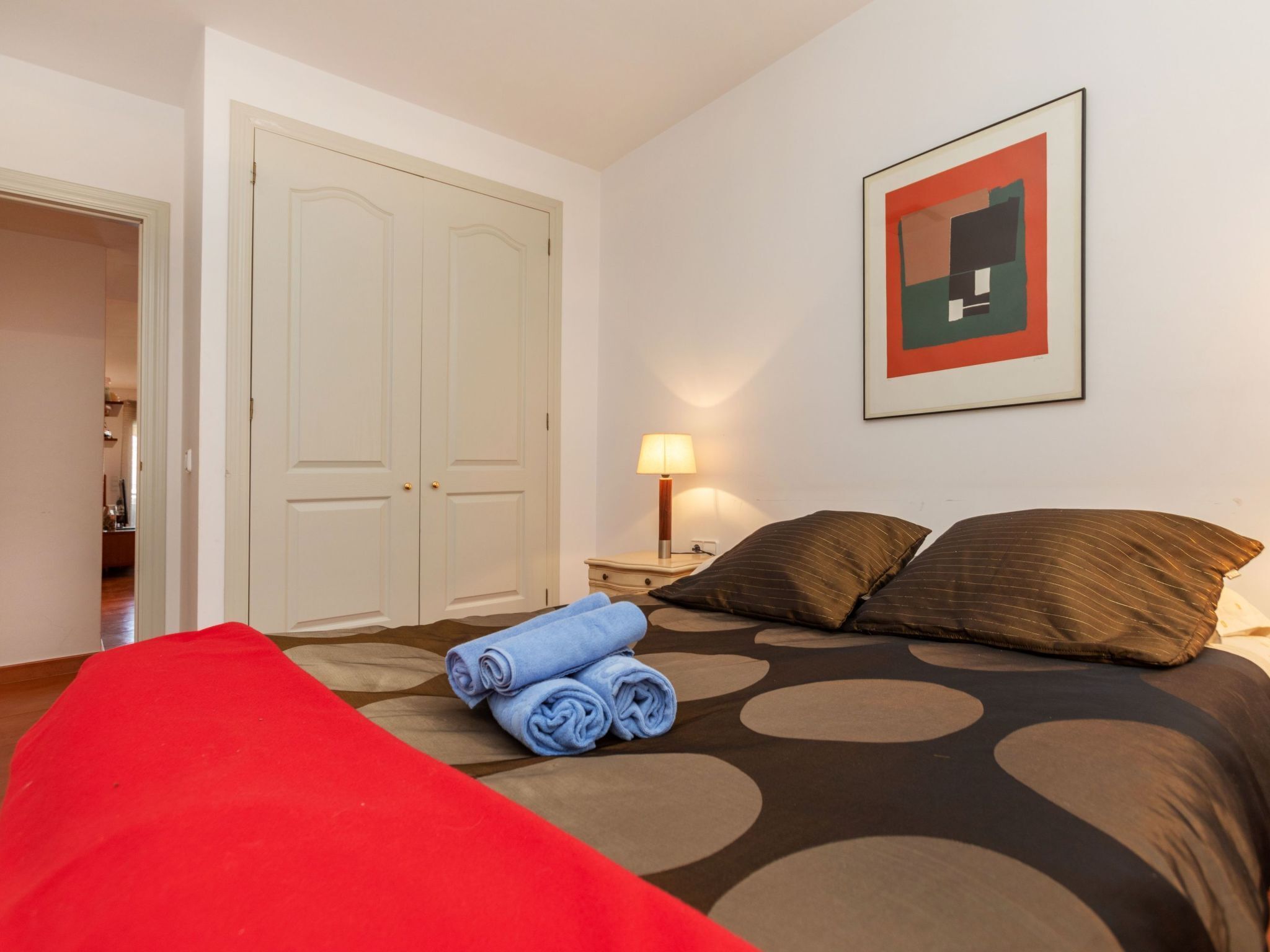 A hotel room with a bed, a red chair, a door, a nightstand with a lamp, and a framed picture on the wall.