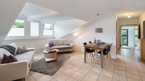 Ein modernes Wohnzimmer mit Sofa, Couchtisch, Esstisch und Stühlen. Es gibt Fenster mit Jalousien, eine Lampe und einen Blick ins Grüne.