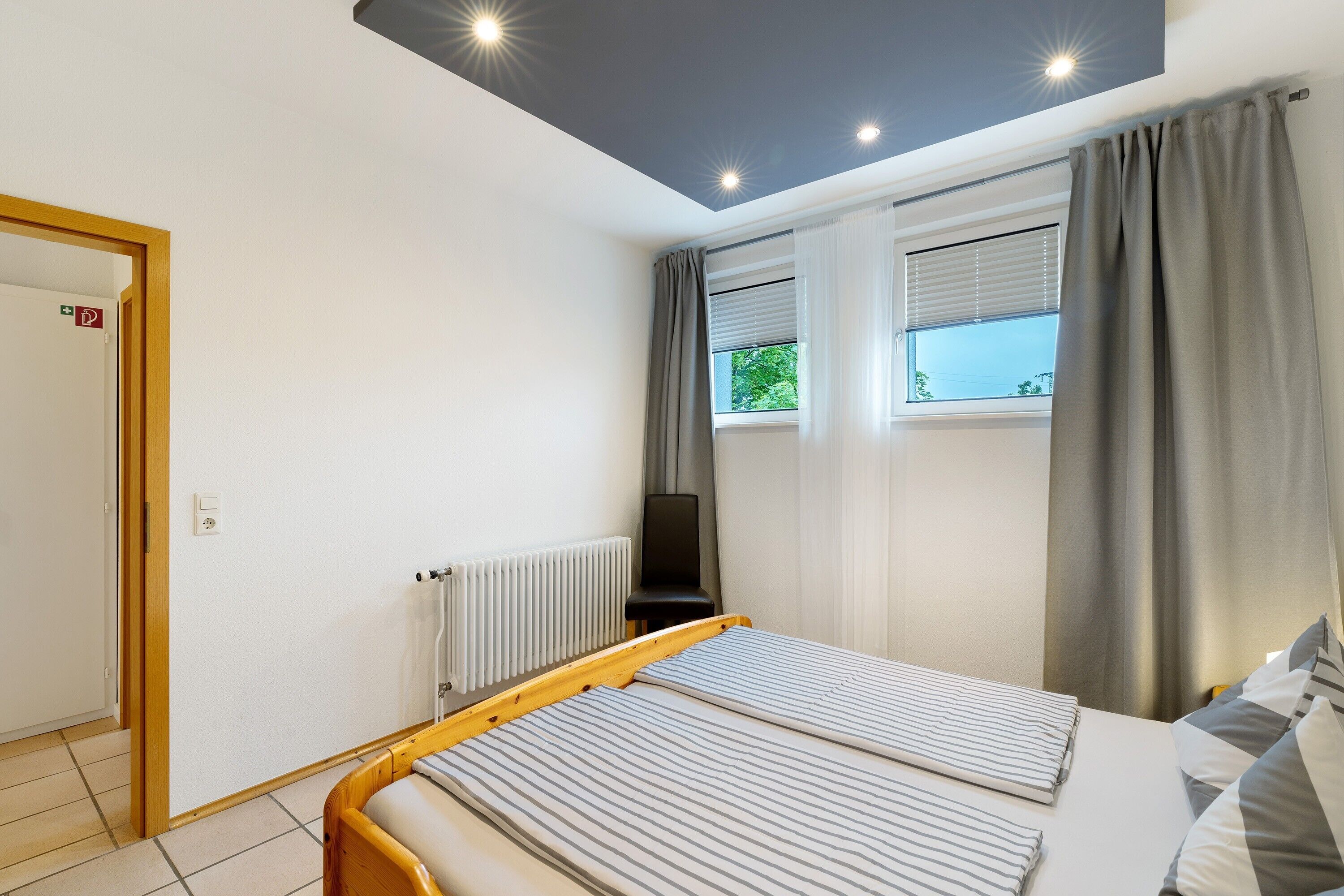 Quarto com cama, radiador, janela com cortinas e porta com placa de saída de emergência.