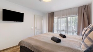 Une chambre à coucher avec un lit, un téléviseur à écran plat fixé au mur, une fenêtre avec des rideaux légers et une vue sur l’extérieur.