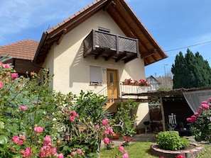 Une maison avec un balcon, un toit en bois et un jardin où poussent des fleurs roses.