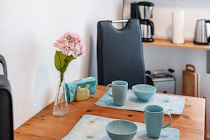 Une table à manger ornée d’un vase de fleurs, avec une machine à café et une bouilloire posées sur une étagère.