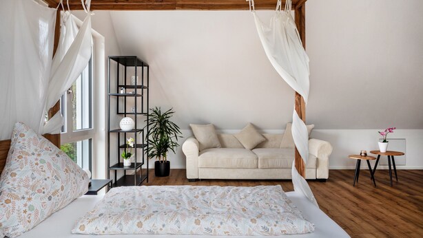 Een slaapkamer met een hemelbed, een bank, een klein tafeltje, een kamerplant en een boekenkast.