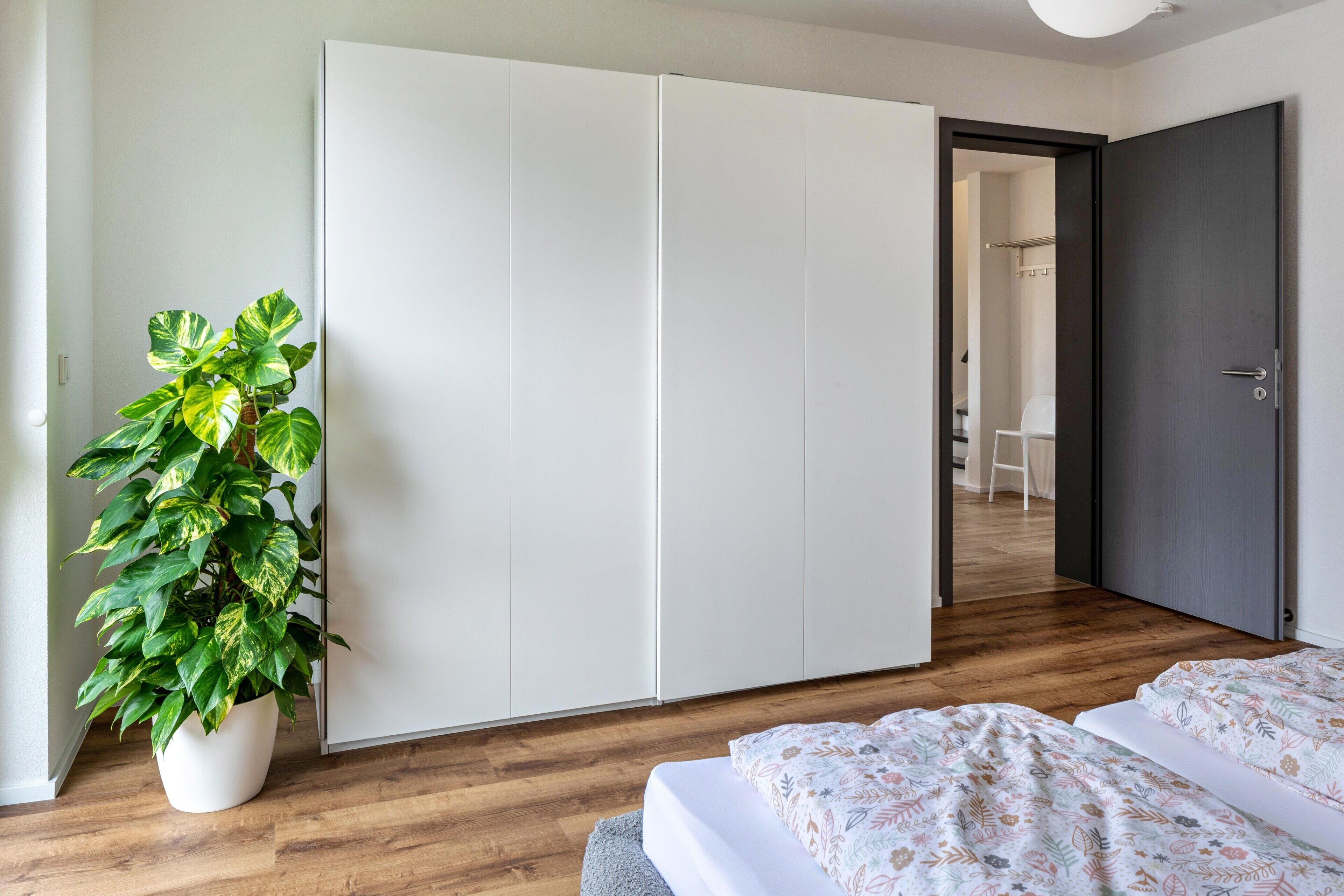 A bedroom with a bed, a wardrobe, a potted plant, and a door leading to another room.