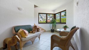 Habitación de esquina con un sofá verde, sillas de mimbre, una mesita con una planta y vistas a casas a través de una ventana.
