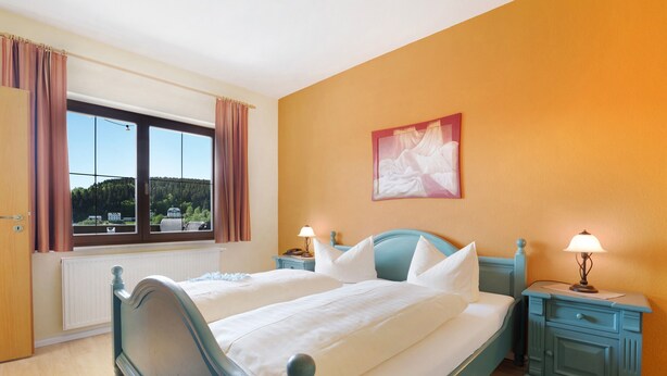 A double bed with a headboard, two bedside tables, a window with curtains, a radiator, and a painting on the wall.