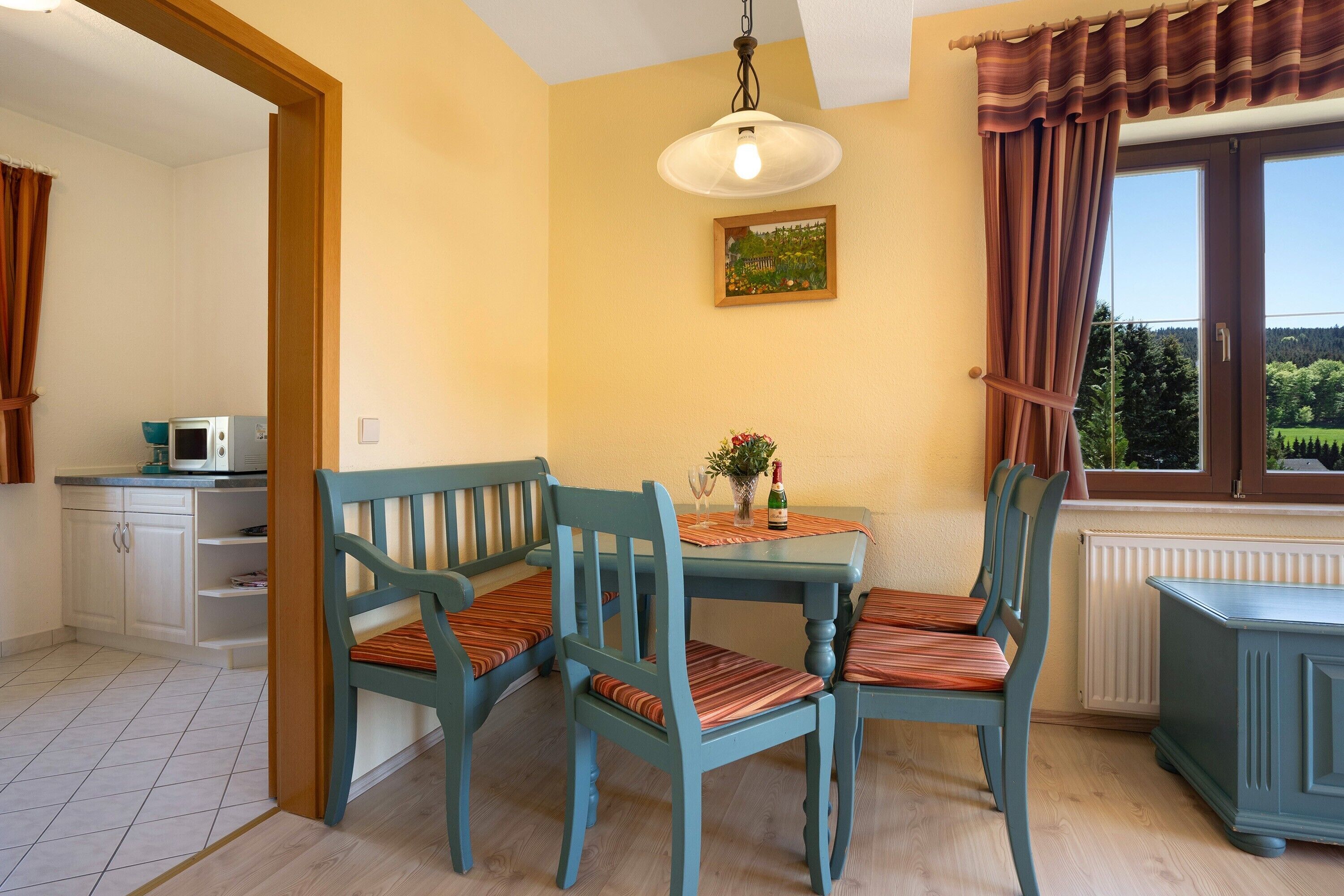A dining area with a table and chairs, a hanging light fixture, a window with curtains, a radiator, and a microwave in the background.