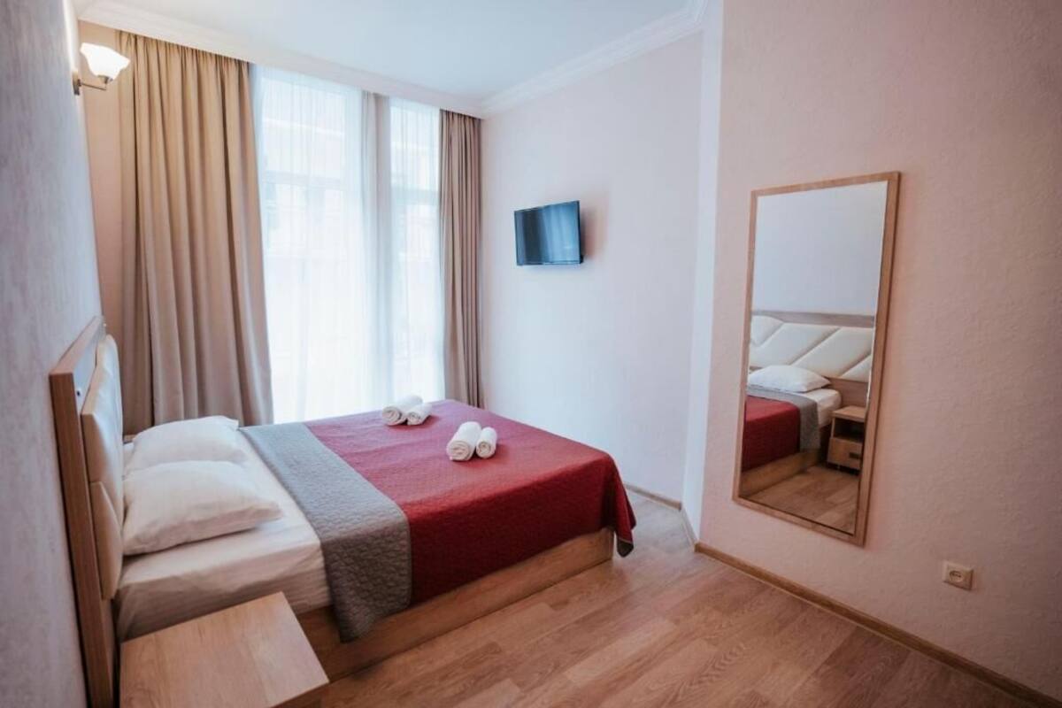 A hotel room with a bed, a TV, a mirror, and a window with curtains.