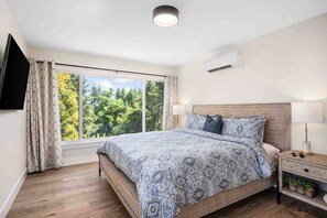 A bedroom with a large window, a bed with patterned bedding, a bedside table with a lamp, and a small shelf with plants. - Breathtaking Wine Country Home w/ Incredible Dundee Hills Vineyard Views, Covered Deck w/ Hot Tub (Dundee)