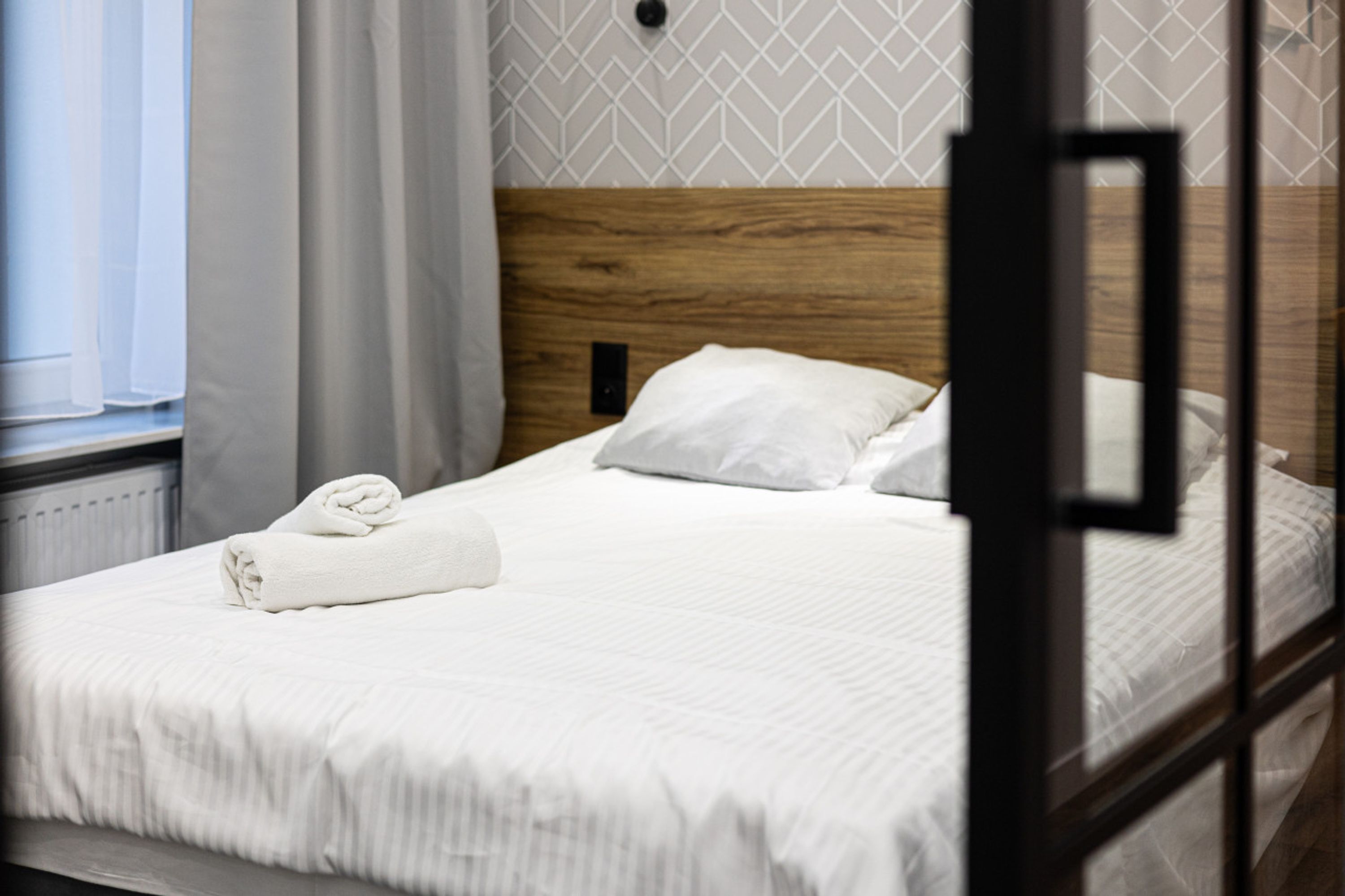 Una cama bien hecha con sábanas blancas y una toalla enrollada, un cabecero de madera y una ventana con cortinas.