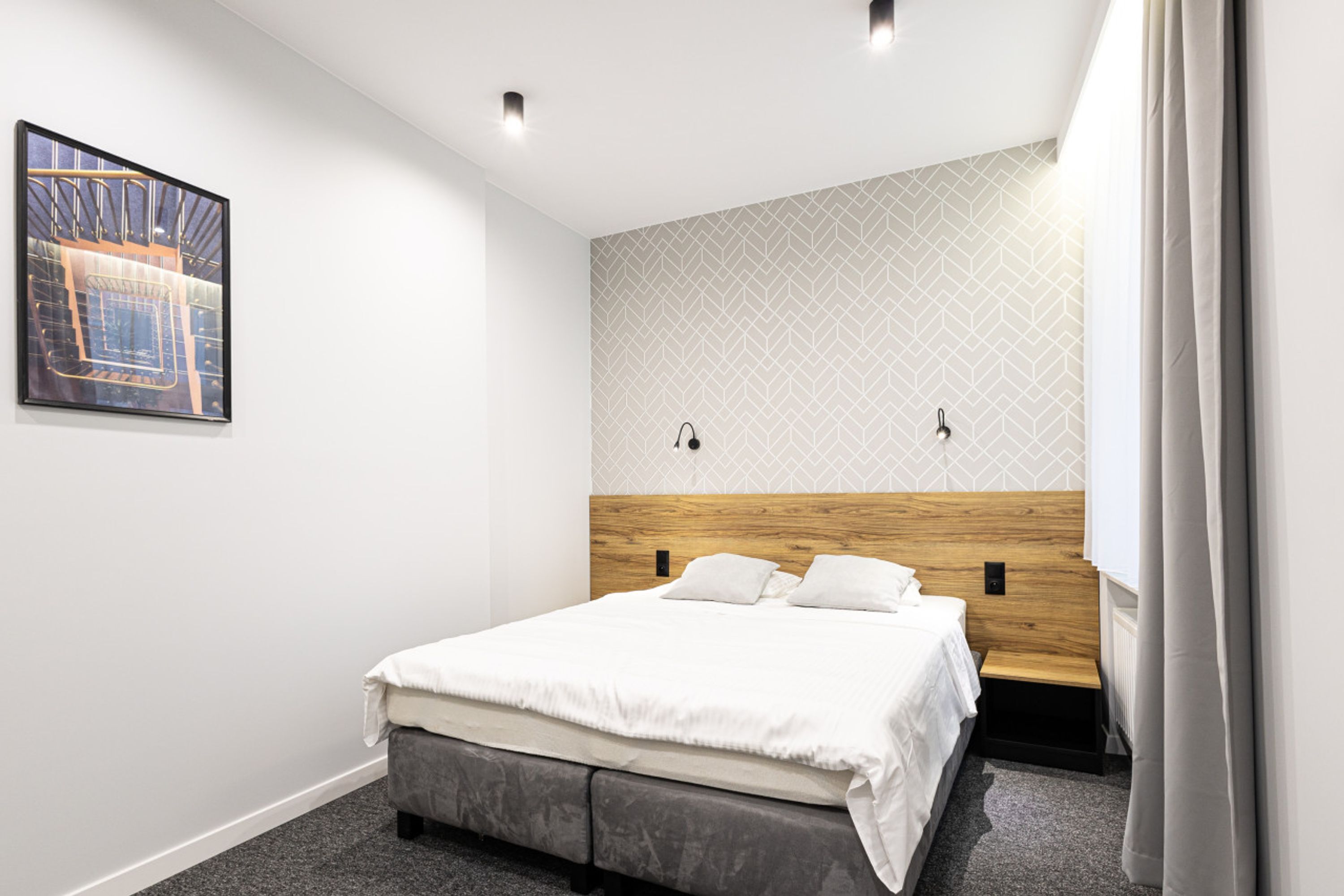 Un dormitorio moderno con cabecera de madera, cama grande, pequeño banco y pared con estampado geométrico.