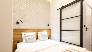 Una habitación de hotel moderna con cabecera de madera, una cama grande con ropa blanca, un armario con puerta corredera de vidrio y una pared con estampado geométrico.