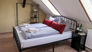 Un dormitorio con techo inclinado, una cama con sábanas blancas y almohadas rojas, una mesita de noche con una botella y vasos, y un radiador.