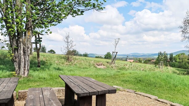 Un área de picnic con bancos de madera y suelo de grava, rodeada de campos cubiertos de hierba y árboles.