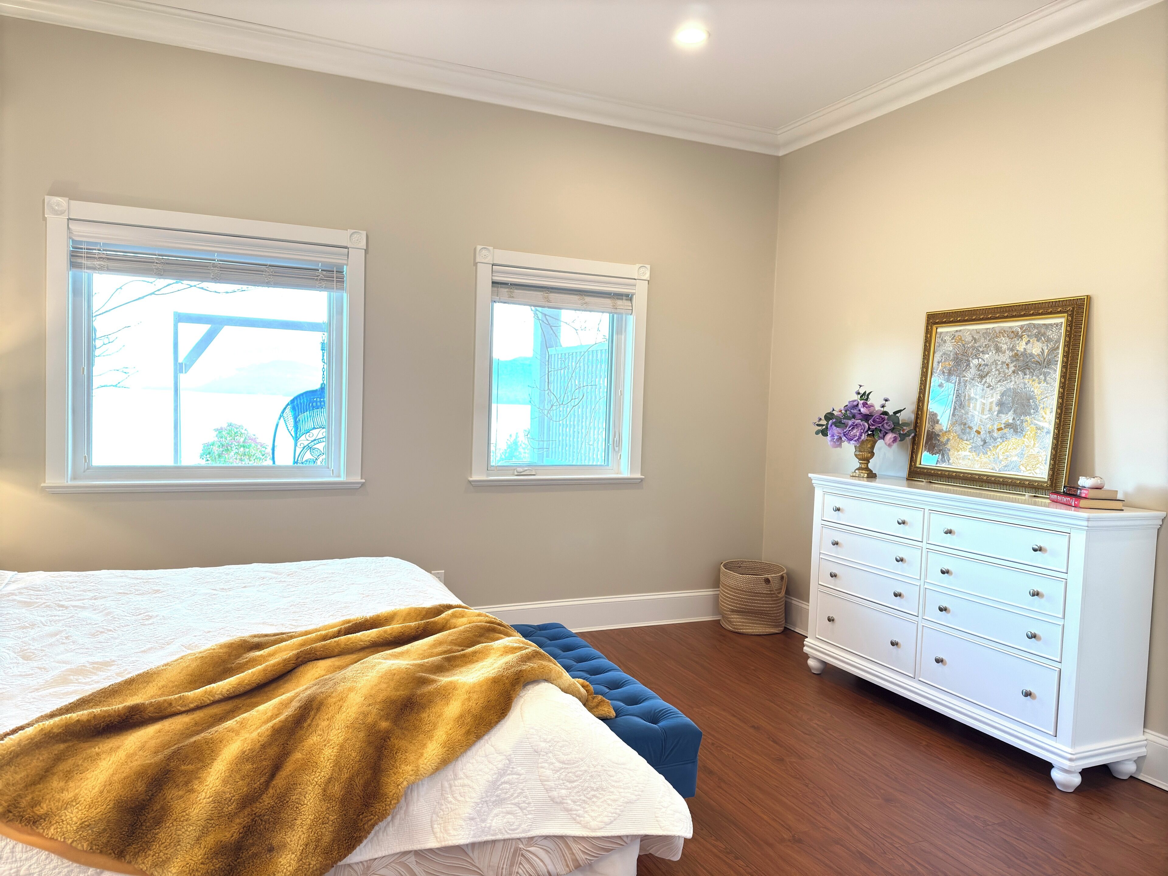 Une chambre à coucher avec un grand lit, deux fenêtres, une commode et un tableau encadré posé sur la commode.