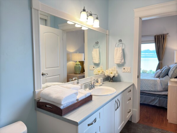 Un baño con un espejo grande, un lavabo blanco y un mueble con cajones. Hay un perchero sobre el espejo y una ventana con cortinas.