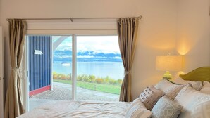 A bedroom with a bed, pillows, a nightstand with a lamp, and a view of a lake through a sliding glass door. - Blue House (Gibsons)