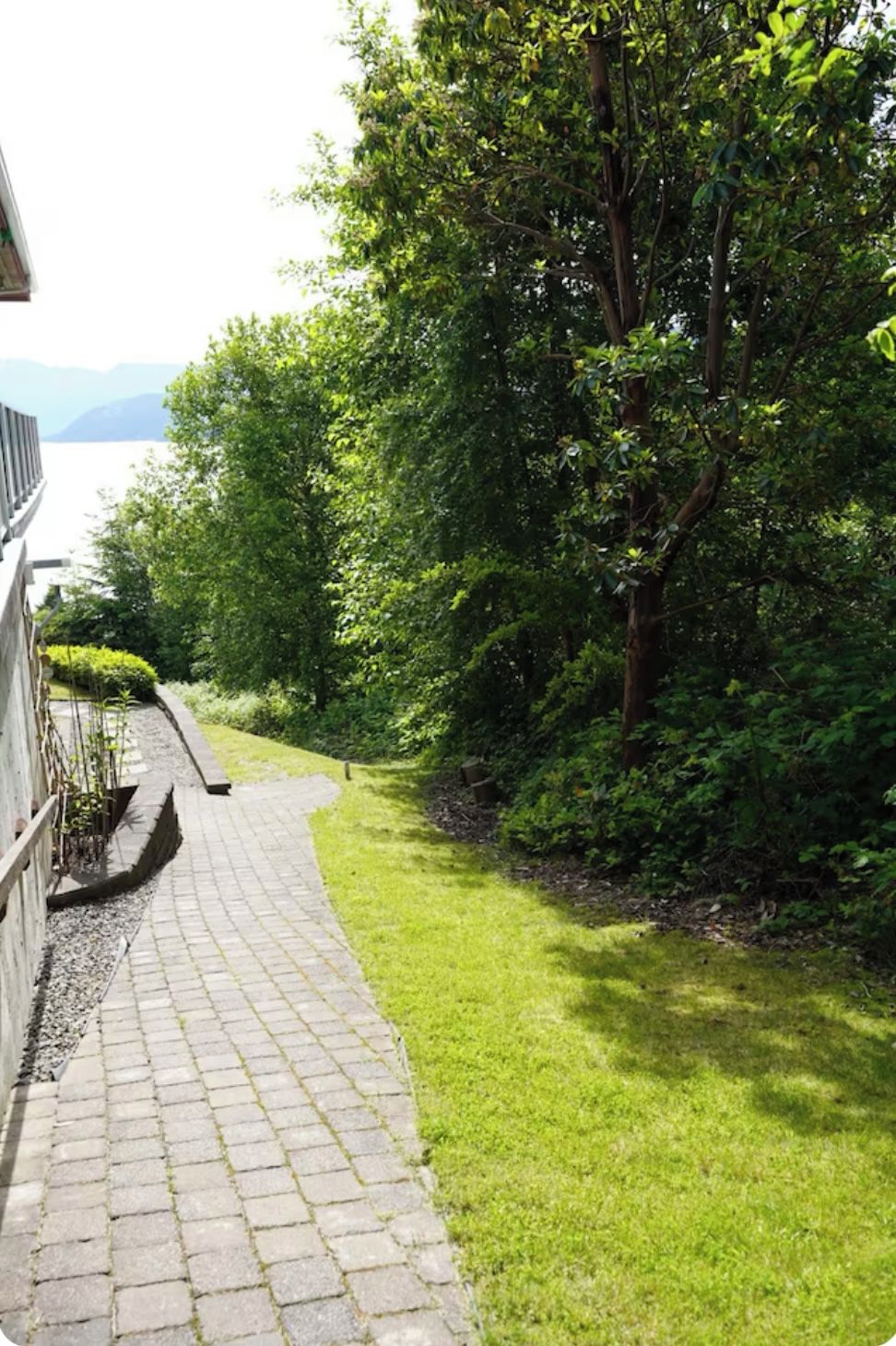 A paved pathway leading through a lush green garden with dense foliage and a view of a body of water in the distance.