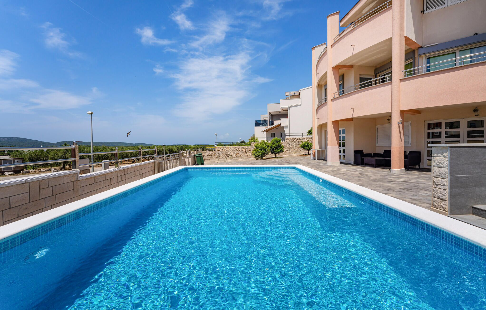 A swimming pool with clear blue water, surrounded by a stone deck and a building with balconies.
