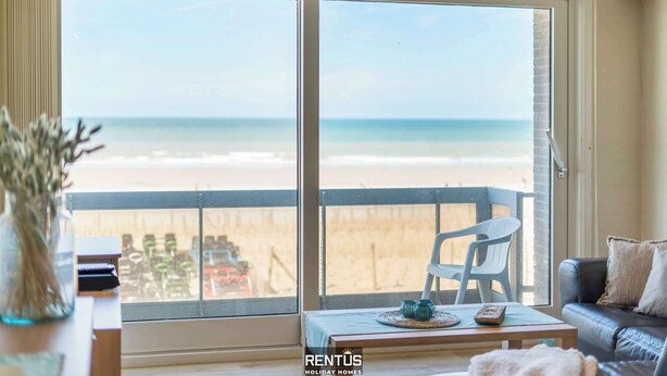Ein Zimmer mit einer großen Schiebeglasdoor, die auf einen Strand blickt. Ein weißer Stuhl und ein kleiner Tisch mit Fernbedienung und Dekorationen sind ebenfalls vorhanden.