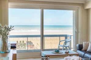 Habitación con un amplio ventanal corredero que da a una vista de la playa. Se observa una silla blanca y una mesita con un control remoto y objetos decorativos.