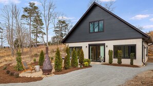 Une maison moderne à deux étages, dotée d’un toit foncé, de grandes fenêtres et d’un jardin bien entretenu agrémenté de divers arbustes et d’une structure métallique décorative.