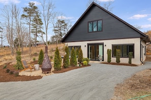 A modern two-story house with a dark roof, large windows, and a well-maintained garden with various shrubs and a decorative metal structure.