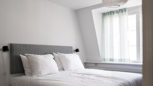Un dormitorio con cama, ropa de cama blanca, cabecera gris, ventana con cortinas translúcidas y una lámpara de pared.