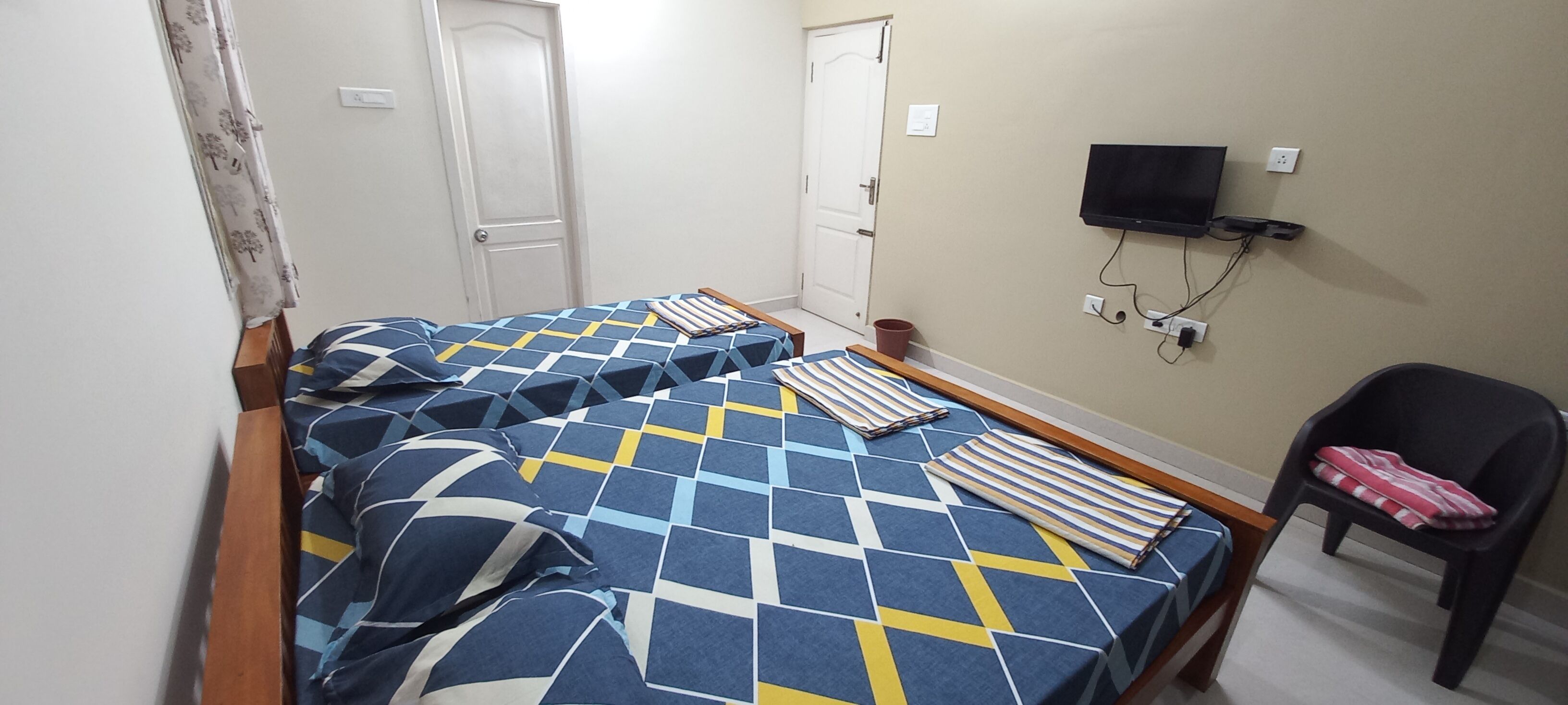 A room with two beds, a television mounted on the wall, and a chair.