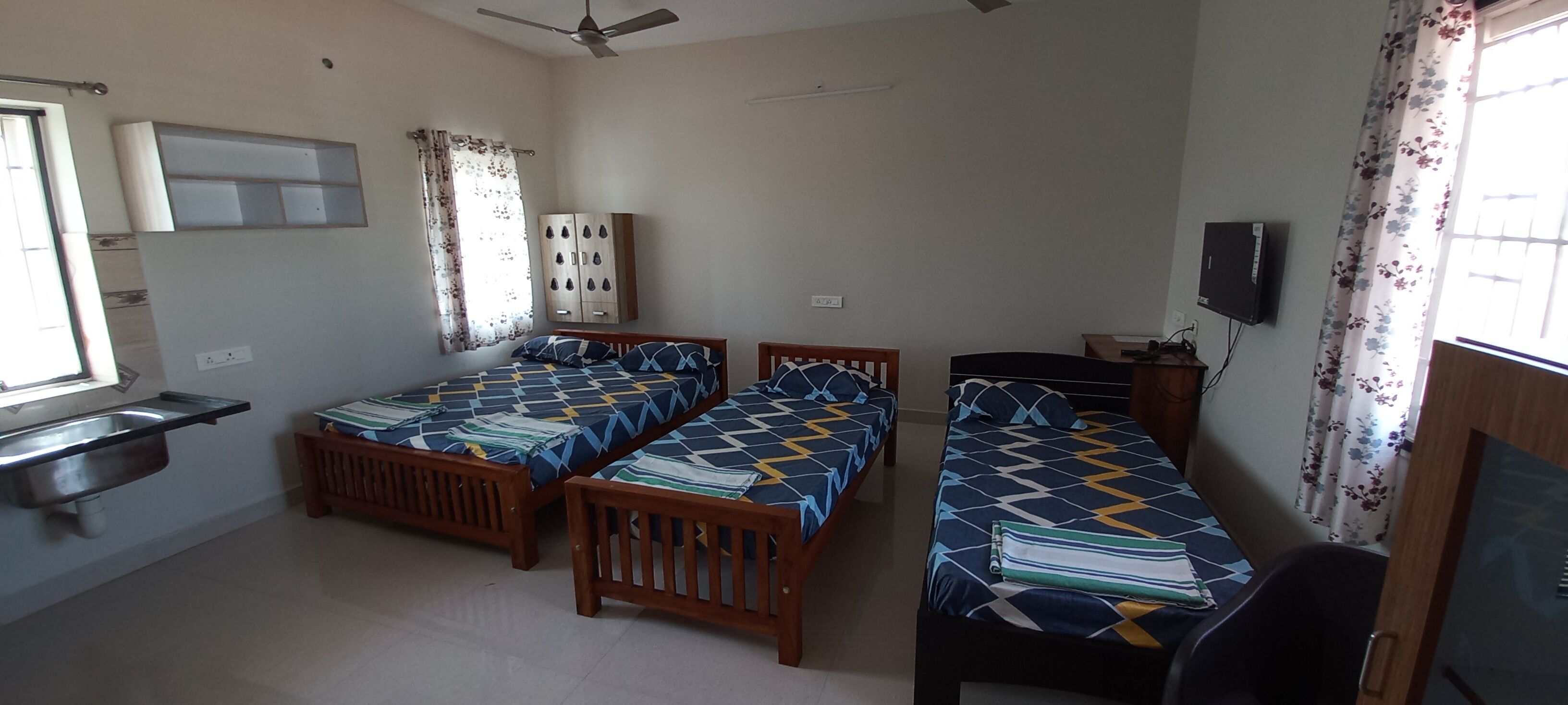 A room with two wooden beds, a small desk, a ceiling fan, a TV, and a window with curtains.