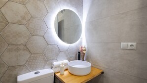 Ein Badezimmer mit Wand aus sechseckigen Fliesen, einem runden Spiegel, einem Holzwaschtisch, einer Toilette und einem Waschbecken mit runder Schüssel.