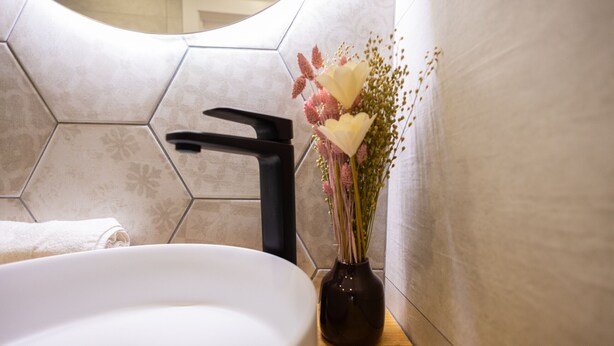 Une salle de bain avec un lavabo blanc, un robinet noir et un vase rempli de fleurs posé sur un comptoir en bois.