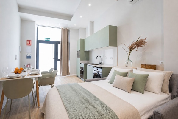 A modern hotel room with a bed, a dining area, and a kitchenette.