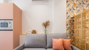 A grey sofa with two orange cushions, a microwave, a vase with dried plants, and a wall with a tropical leaf pattern.