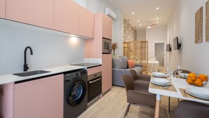 A modern kitchen with a washing machine, a dining area with a table set for two, and a living area with a sofa and a patterned wall.