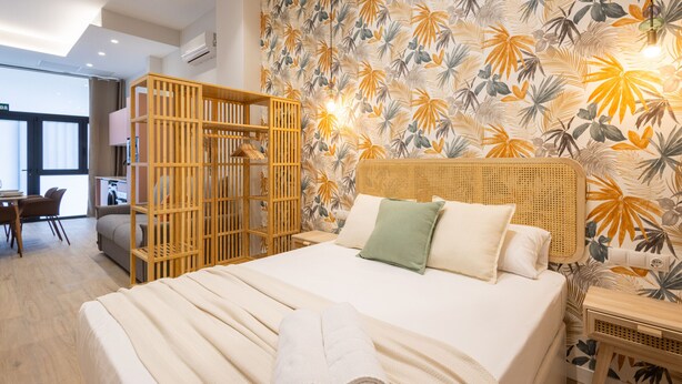 A bedroom with a bed, a wooden headboard, a wall with a tropical leaf pattern, and a wooden wardrobe.