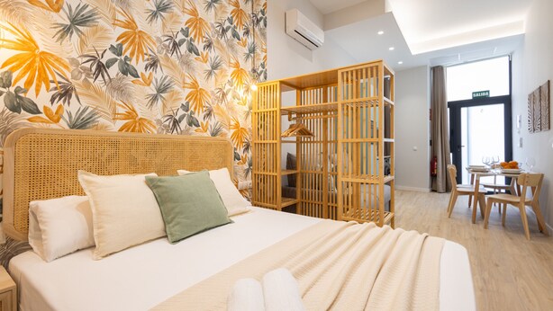 Ein Hotelzimmer mit Bett, Holz-Kopfteil, Wand mit tropischem Blattmuster, einer Holzwand, einem Essbereich mit Tisch und Stühlen sowie einer Klimaanlage.