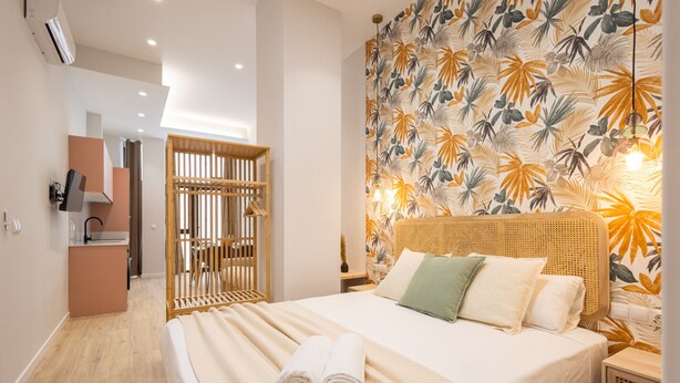 Une chambre d’hôtel avec un grand lit, une tête de lit en bois, un mur décoré d’un motif de feuilles tropicales et une petite table de chevet avec une lampe.