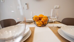 Une table à manger avec un panier en fil métallique contenant des oranges, deux verres à vin et des assiettes blanches.