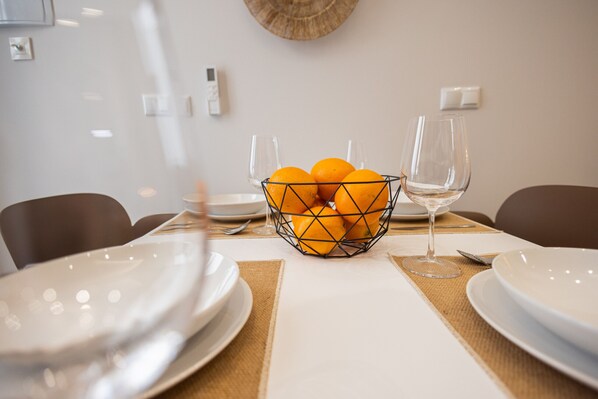 Une table à manger avec un panier en fil métallique contenant des oranges, deux verres à vin et des assiettes blanches.