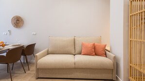 Un canapé aux couleurs neutres agrémenté de deux coussins, une table à manger avec des chaises, et une assiette décorative fixée au mur.