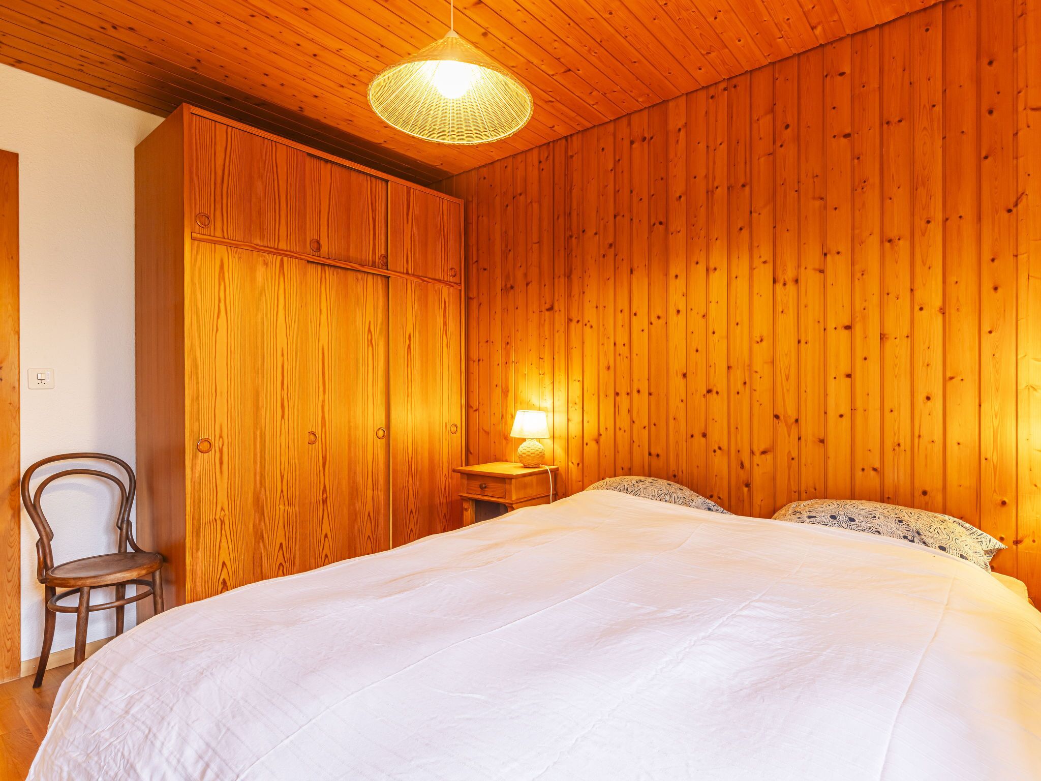 Un dormitorio con paredes de madera, una cama con ropa blanca, una silla y una lámpara en una mesita de noche.