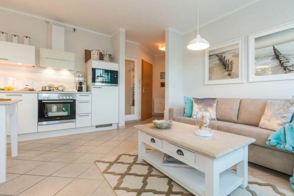 Une cuisine moderne avec des meubles blancs, un four encastré et une machine à café. On y trouve un canapé beige avec un plaid bleu et une table basse blanche agrémentée d’un objet décoratif.