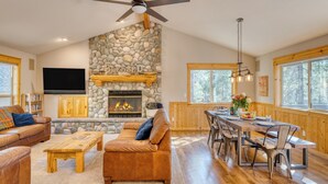 A spacious living room with a stone fireplace, a ceiling fan, and a dining area with wooden furniture. - Quail by AvantStay | Great Location + Game Room, Hot Tub, & Deck! (Sunriver)