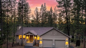 A two-story house with a garage, surrounded by tall pine trees, at dusk. - Quail by AvantStay | Great Location + Game Room, Hot Tub, & Deck! (Sunriver)