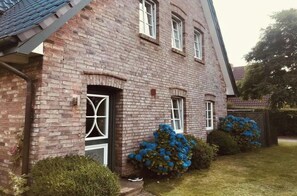 Una casa de ladrillo con puerta principal, dos ventanas y un pequeño jardín con flores azules.