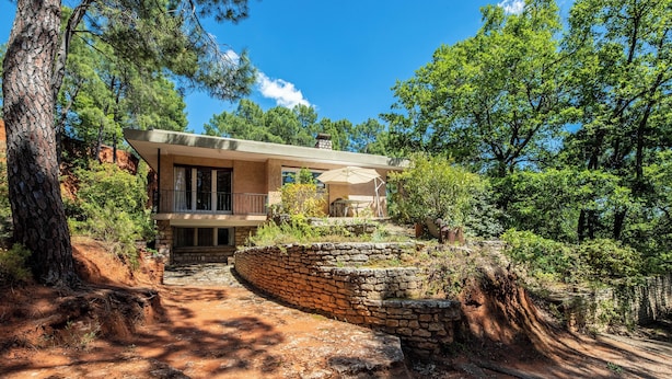 Una casa moderna con una escalera de piedra que lleva a una terraza de madera, rodeada de árboles y un cielo despejado.