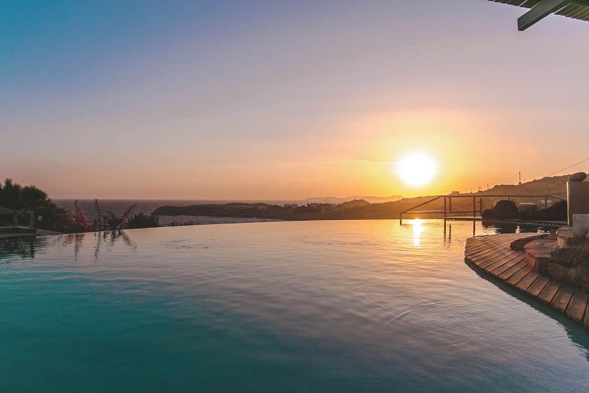A swimming pool at sunset with a wooden deck and a view of the ocean.